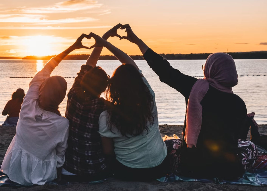 Four women sitting by the shore at golden hour forming hearts with their hands, celebrating friendship, confidence, and sisterhood in a vibrant, sassy vibe.